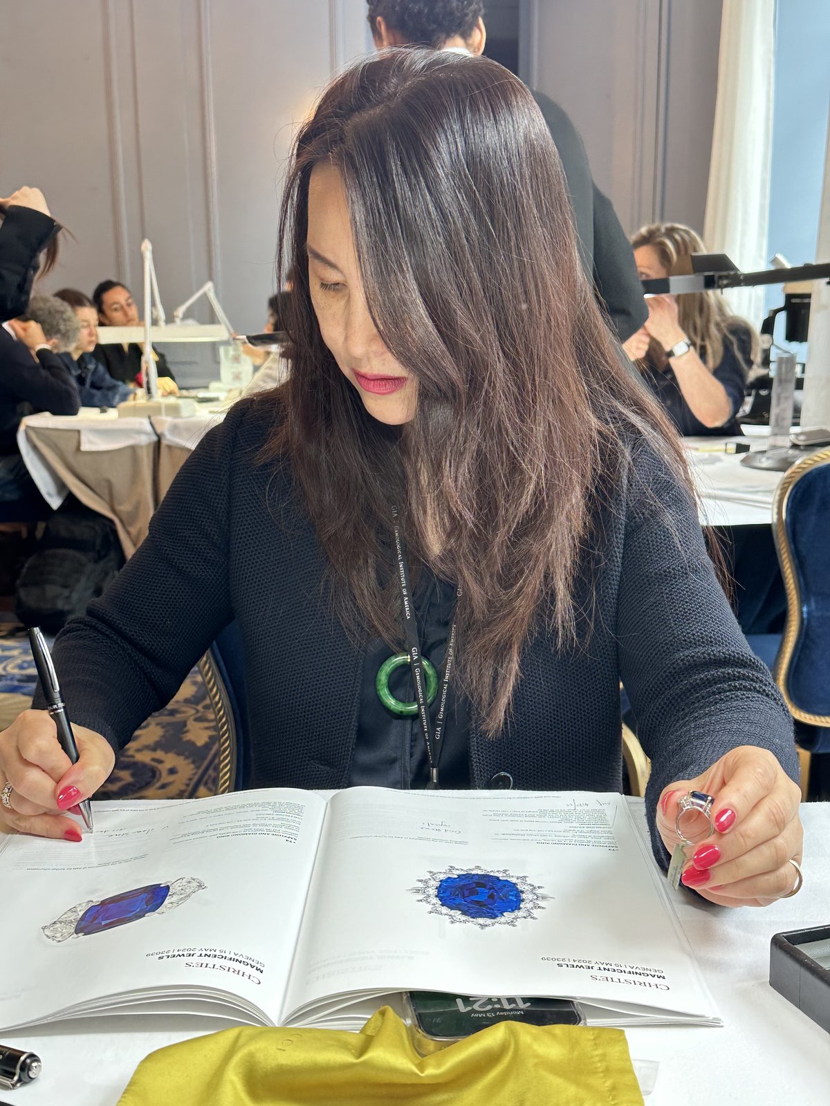 Kehan Li at a Magnificent Jewels auction preview, taking notes against a catalogue showing a sapphire and emerald lot, wearing a green jadeite bangle.